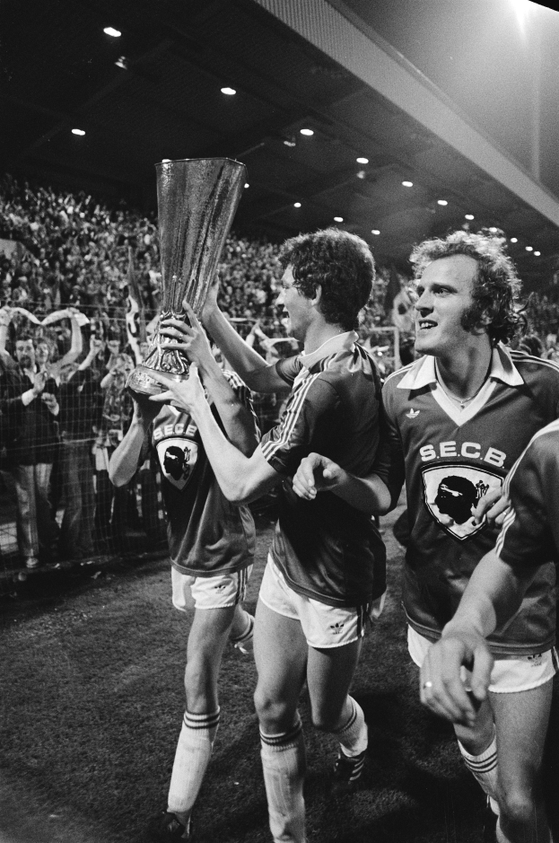 Eine Gruppe von Männern, die einen Pokal auf einem Fußballfeld halten und ihren FA-Cup-Sieg feiern, umgeben von einer Menge hinter einem Zaun, unter Stadionlichtern, in Schwarz-Weiß.