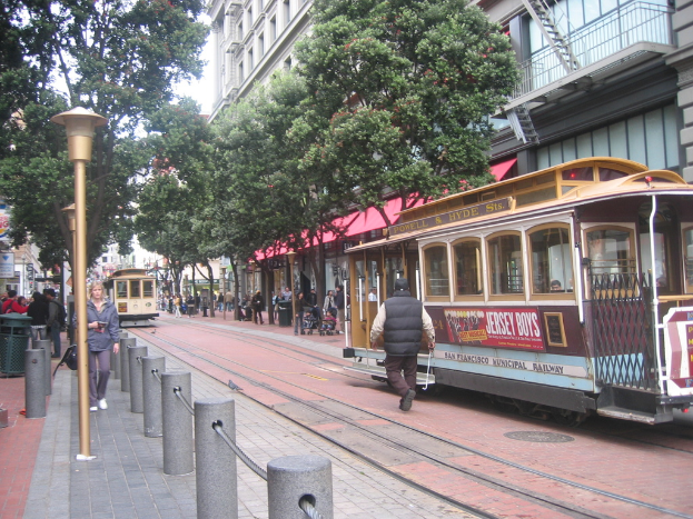 Ein Zug auf Eisenbahnschienen mit Text an seiner Seite, eine Person in der Nähe, kleine Pfosten mit Seilen auf der linken Seite, Straßenlaternen, Bäume, viele Menschen, ein Möbel auf der linken Seite und Gebäude mit Leitern auf der rechten Seite.