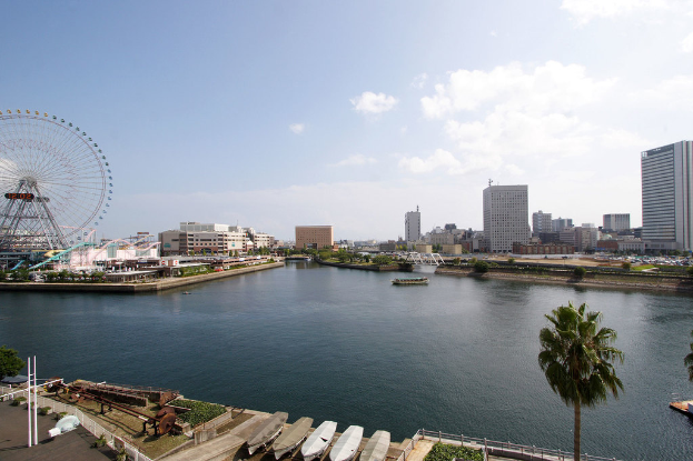 Eine Stadtlandschaft mit Hochhäusern, einem Riesenrad, Vergnügungsanlagen, Schiffen auf dem Wasser und verschiedenen Strukturen wie Geländern, Skulpturen und Absperrpollern, alles unter einem bewölkten Himmel mit Bäumen und Büschen im Vordergrund.
