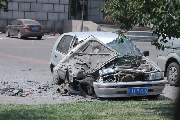 Ein verunglücktes Auto am Straßenrand, umgeben von Gras und einem Baum auf der rechten Seite, mit einem Gebäude im Hintergrund.