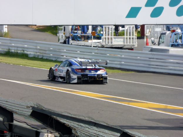 Ein BMW M6 GT3 fährt auf einer Rennstrecke bei den 24 Stunden von Le Mans, umgeben von einer Absperrung, Gras, Verkehrskegeln, Menschen und anderen Gegenständen, sowie Bäumen und einem klaren blauen Himmel im Hintergrund.