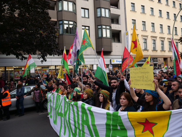 Eine große Gruppe von Menschen marschiert mit Flaggen und Schildern mit Text die Straße entlang, mit einem geparkten Auto auf der rechten Seite und einem Baum auf der linken Seite, vor Gebäuden mit Fenstern und Schildern, was auf eine algerische Demonstration hinweist.