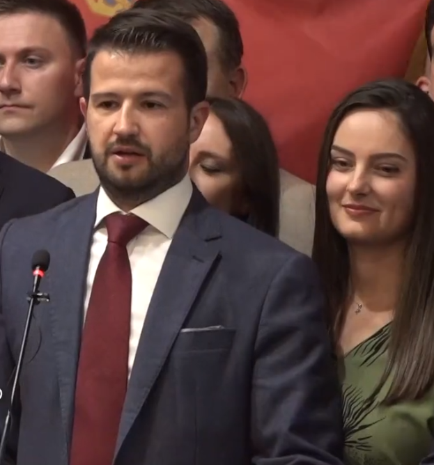 Ein Mann im Anzug und Krawatte steht an einem Podium mit einem Mikrofon und blickt auf eine Menge mit einem Banner im Hintergrund.