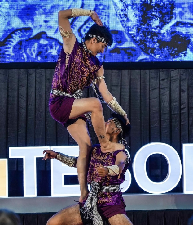 Zwei Performer in violetten Kleidern führen einen akrobatischen Hebemanöver auf der Bühne vor einem Publikum aus