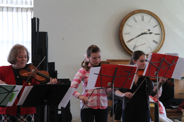 Drei Frauen auf der Bühne mit Violinen stehend und Musikständer vor sich, während eine Gruppe von sitzenden Frauen im Vordergrund zuhört; eine Uhr hängt an der weißen Wand im Hintergrund, mit einem weißen Vorhang in der linken Ecke.