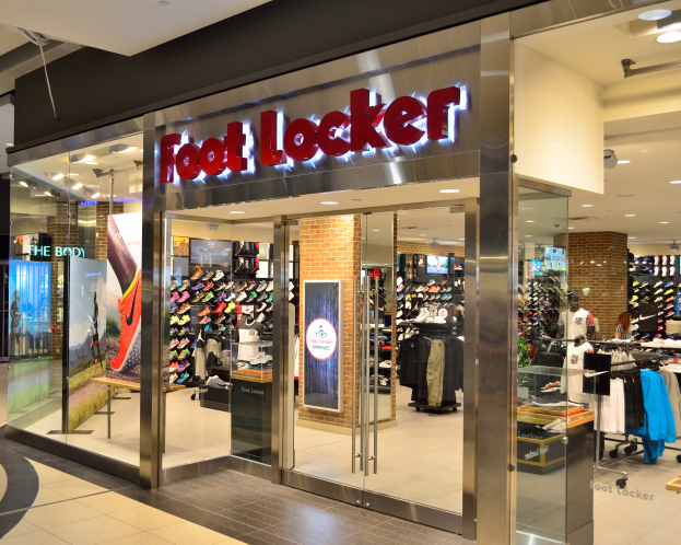 Ein Foot Locker Store in einem Einkaufszentrum mit Glaswänden und -türen, Kleider an Bügeln, Schaufensterpuppen in Kleidern, Informationsschilder, Deckenleuchten und verschiedene andere Gegenstände.