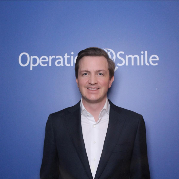 Ein Mann im Anzug lächelt stolz vor einer blauen Wand mit der Aufschrift "Operationsmile".