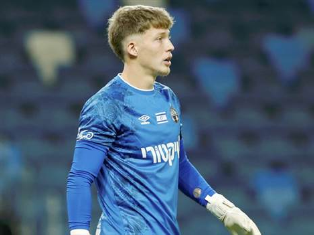 Ein junger Mann in einem blauen Torwart-Outfit und weißen Handschuhen steht auf einem Fußballfeld während eines Spiels, mit einem unscharfen Hintergrund.