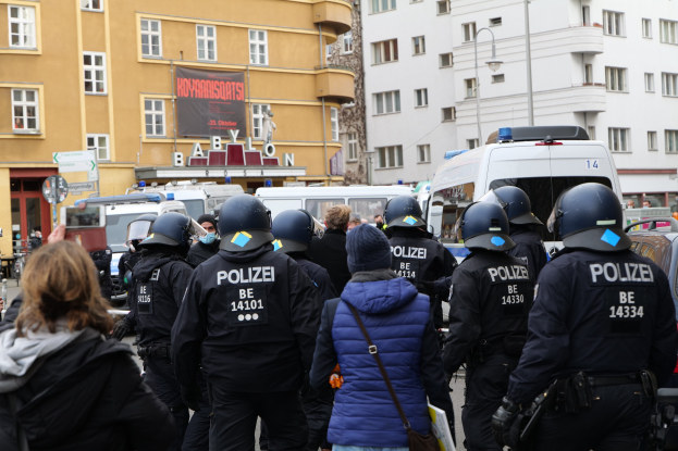 Polizeibeamte in Uniform stehen vor einer Menge mit Helmen und Jacken während einer Demonstration in Berlin, Deutschland, mit Fahrzeugen, Gebäuden, Laternenmasten und einem Banner im Hintergrund.