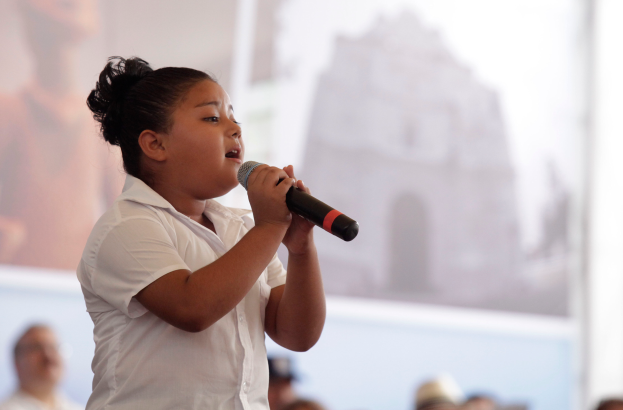 Ein junges Mädchen singt in ein Mikrofon vor einer Menge, mit einem Bildschirm im Hintergrund.