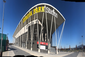 Großes Stadiongebäude mit Text, umgeben von Pfählen, Lichtern, Bäumen und einem Zaun, unter einem sichtbaren Himmel.