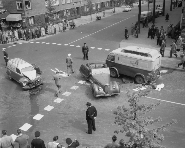 Schwarz-weiß-Szene eines Autounfalls an einer Straßenkreuzung mit mehreren Fahrzeugen, darunter ein Lieferwagen, und einer Gruppe von Schaulustigen; Gebäude, Bäume, Pfosten und Texttafeln sind im Hintergrund sichtbar.