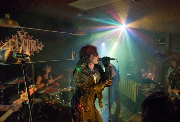 Eine Frau in einer schwarzen Jacke singt auf der B├╝hne in ein Mikrofon, hinter ihr spielt ein Mann mit Sonnenbrille und Stirnbinde ein Instrument; im Hintergrund sind ein "LA Guns"-Plakat und Diskoleuchten zu sehen.