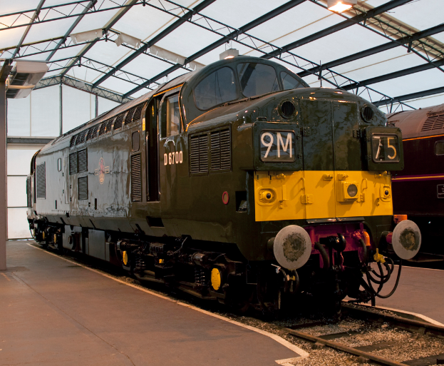 Ein Zug fährt auf den Schienen neben einer Plattform mit einem am Dach befestigten Mast, der Metallstangen mit Lampen trägt.