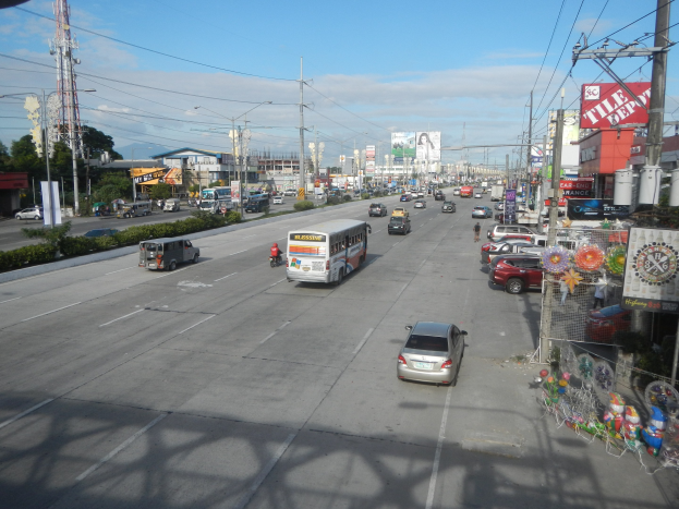 Eine belebte Stadtstraße mit Autos, Bussen, Lastwagen, Strommasten, Gebäuden mit Schildern, Bäumen, Pflanzen, einem Turm im Hintergrund und einem bewölkten Himmel.
