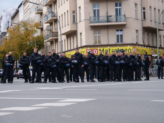 Eine Gruppe von Polizisten in schwarzen Uniformen mit blauen Helmen steht in einer Straße, die von Laternen, Bäumen und Glasfenstergebäuden gesäumt ist, unter einem klaren blauen Himmel.