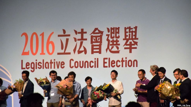 Eine Gruppe von Menschen auf einer Bühne mit Blumensträußen, darunter ein Publikum und ein Bildschirm mit Text und Logo für die Wahl des Legislativrats 2016.