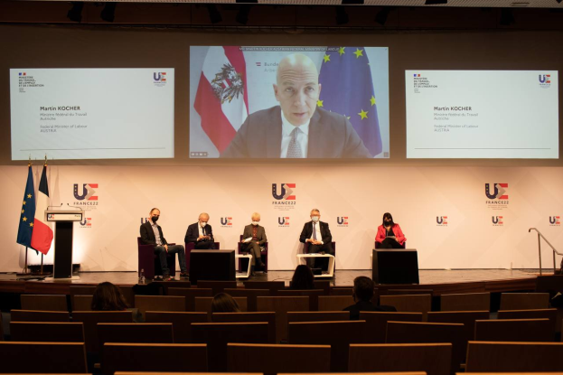 Eine Gruppe von Menschen auf einer Bühne in einem halben Kreis um ein zentrales Podium mit Mikrofon, flankiert von Flaggen, mit einem Bildschirm im Hintergrund, der Text und Bilder anzeigt, während einer Pressekonferenz der Europäischen Union.
