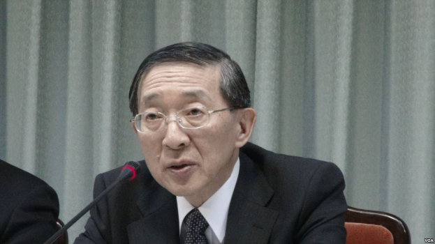 Ein Mann im Anzug und Krawatte mit Brille sitzt an einem Tisch mit Mikrofon, mit einer Hand auf der linken Seite und einem Vorhang im Hintergrund.