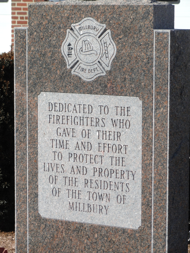 Denkmalstein mit einer Gedenktafel für Feuerwehrleute in Millbury, vor einem Backsteingebäude mit umliegender Vegetation aufgestellt.
