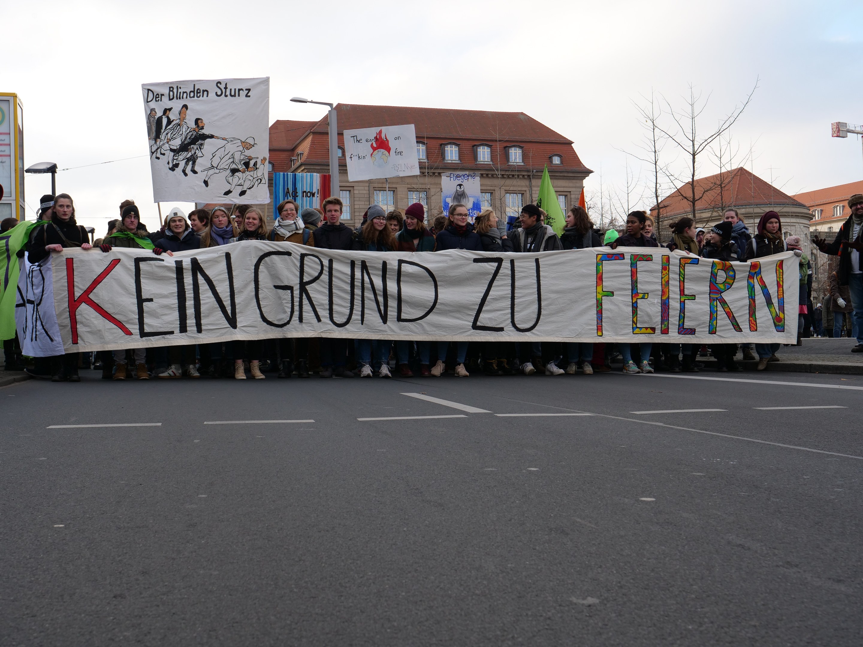 Eine Gruppe von Menschen mit einem Transparent mit der Aufschrift "Kein Grund zu Feiern" protestiert gegen Sparmaßnahmen auf einer Stadtstraße mit Gebäuden, Bäumen und einem klaren Himmel.