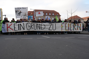 Eine Gruppe von Menschen mit einem Transparent mit der Aufschrift "Kein Grund zu Feiern" protestiert gegen Sparmaßnahmen auf einer Stadtstraße mit Gebäuden, Bäumen und einem klaren Himmel.