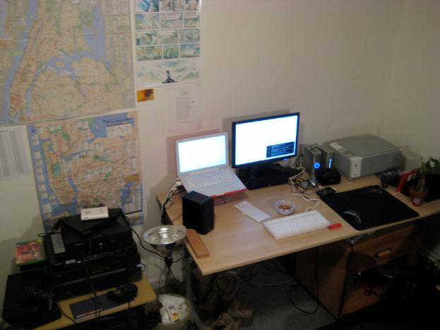 Ein Raum mit einem Tisch, auf dem ein Laptop, Lautsprecher, Monitor, Tastatur, Ball, Maus, Scanner und Papiere liegen, und einem weiteren Tisch mit Elektronikgeräten, mit Charts und Postern an der Wand dahinter.