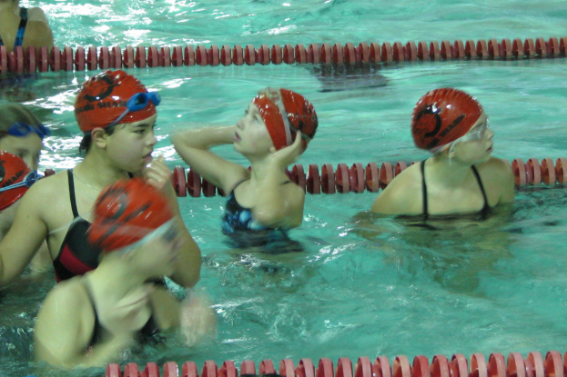 Kinder in einem Schwimmbad mit Badekappen und Schwimmbrillen, mit Wasser im Hintergrund.