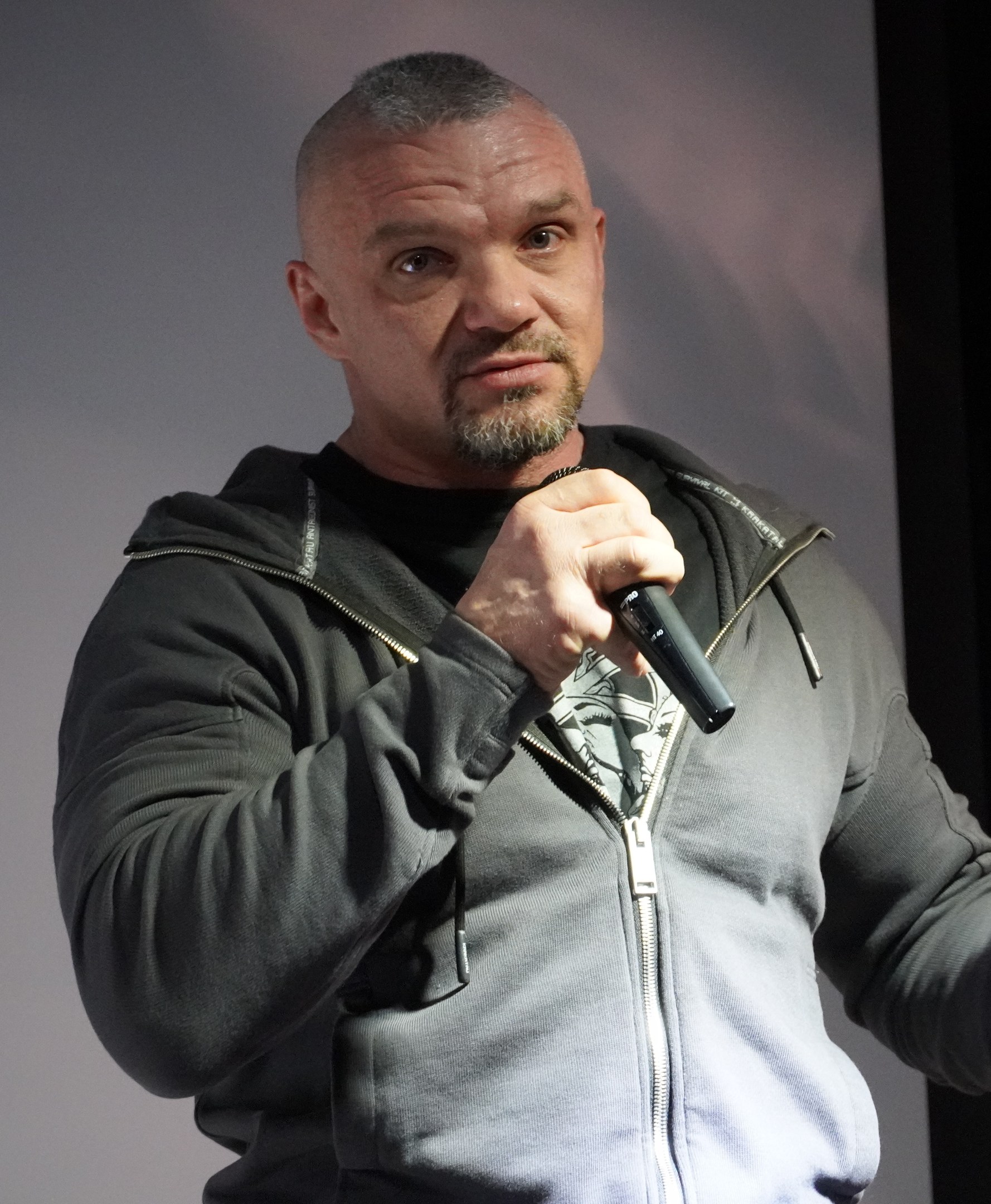 Ein Mann in einer schwarzen Kapuzenjacke und einem schwarzen Anzug mit einem wei├čen Hemd, der ein Mikrofon h├Ąlt, steht selbstbewusst vor einer Wand.