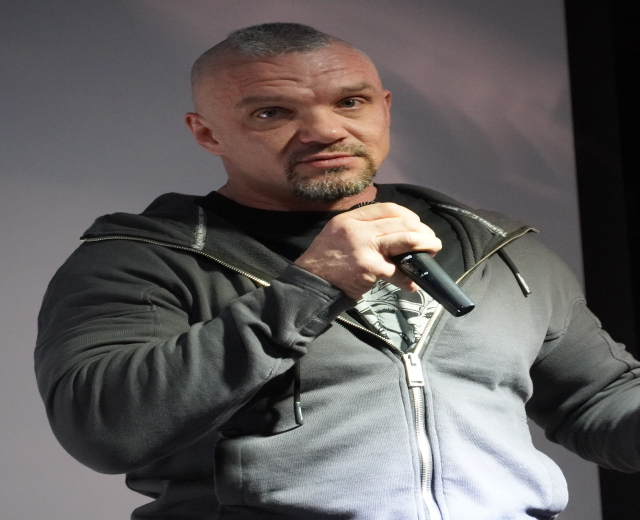 Ein Mann in einer schwarzen Kapuzenjacke und einem schwarzen Anzug mit einem wei├čen Hemd, der ein Mikrofon h├Ąlt, steht selbstbewusst vor einer Wand.