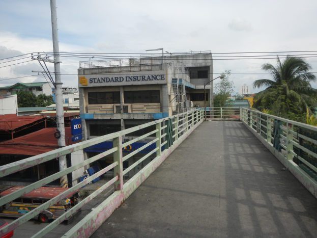 Eine Brücke mit Geländern, Fahrzeuge auf der Straße, Gebäude, Bäume, Strommasten mit Kabeln und einen bewölkten Himmel im Hintergrund, mit einem Gebäude an der Seite mit einer "Standard Insurance"-Aufschrift.