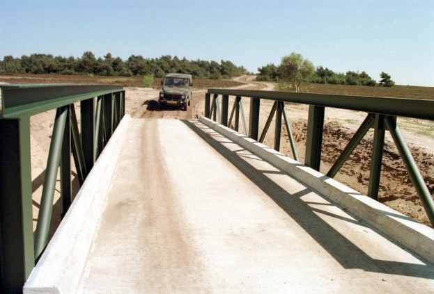Jeep auf einer Brücke über eine Schotterstraße mit Geländern, Bäumen im Hintergrund und einem klaren blauen Himmel.