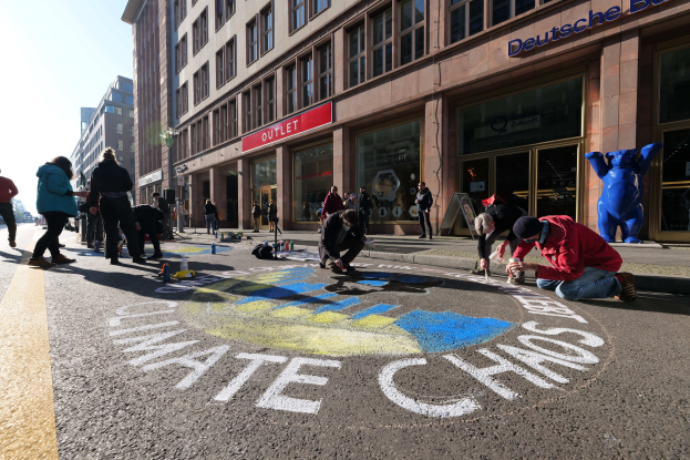 Menschen sitzen vor einem Gebäude mit Fenstern und Namensschildern auf dem Boden, umgeben von Flaschen und anderen Gegenständen, während sie an einer Klimaprotest in Berlin mit Bäumen und einem klaren blauen Himmel teilnehmen.