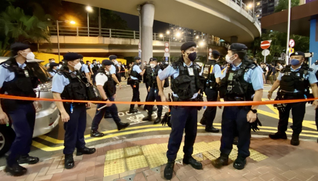 Polizeibeamte in Uniform und mit Masken vor einer Menge mit Fahrzeugen, Gebäuden und einer Brücke im Hintergrund, wahrscheinlich nach einer Demonstration in Hong Kong.