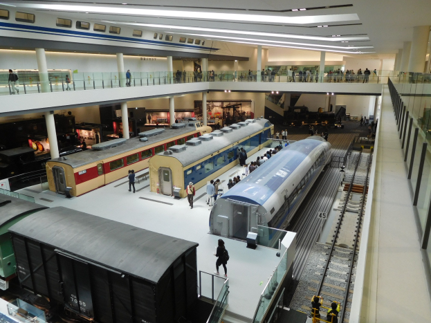 Eine große Museumsausstellung zeigt verschiedene Züge in unterschiedlichen Größen und Farben, mit Menschen, die zuschauen, und Geländern, Säulen und Deckenlampen.