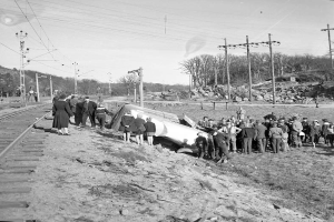Eine Gruppe von Menschen steht um einen Zug herum, der auf einem Schotterhaufen auf einem Bahngleis gelandet ist, mit Strommasten, Drähten, Bäumen und Himmel im Hintergrund, dargestellt in Schwarz-Weiß.