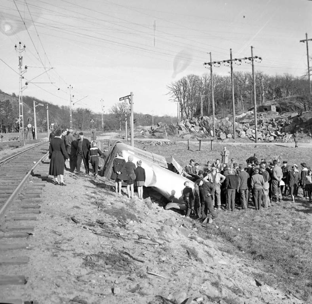Eine Gruppe von Menschen steht um einen Zug herum, der auf einem Schotterhaufen auf einem Bahngleis gelandet ist, mit Strommasten, Drähten, Bäumen und Himmel im Hintergrund, dargestellt in Schwarz-Weiß.