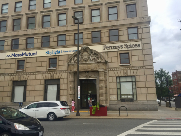 Großes beiges Gebäude mit "Massachusetts Mutual Life Insurance Company"-Schriftzug, Autos vor dem Gebäude geparkt, Straßenlaternen, fahrende Fahrzeuge, Fußgänger auf dem Gehweg, Bäume, Topfpflanzen und bewölkter Himmel.