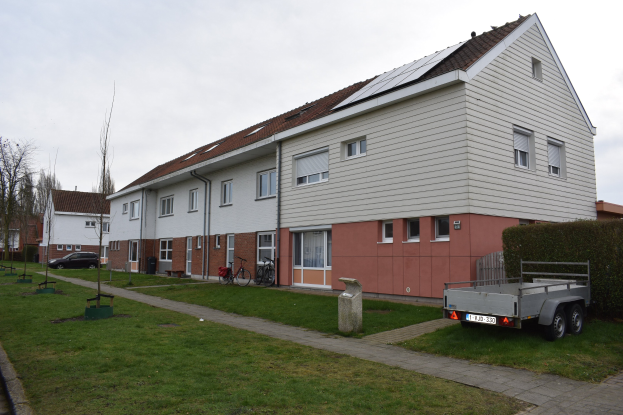 Ein Lastwagen vor einer Reihe von Häusern mit Solarpaneelen auf den Dächern, mit Fenstern, Türen und in der Nähe geparkten Fahrrädern auf einem grasbewachsenen Bereich mit einem Weg, Pflanzen, Bäumen und einem bewölkten Himmel.