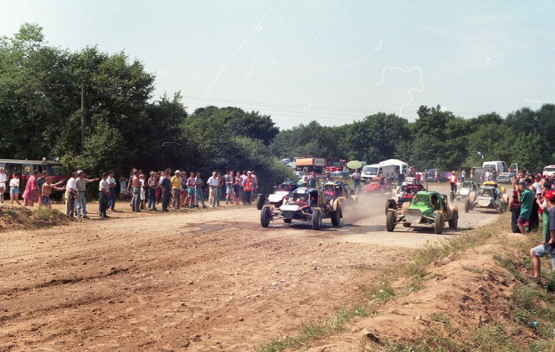 Eine Gruppe von Autos fährt auf einer Schotterstraße mit einer Menschenmenge, einige tragen Mützen, auf einer grasbewachsenen Fläche mit Bäumen und einem klaren blauen Himmel.