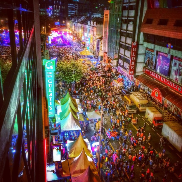 Ein lebhaftes Volksfest-Szenario mit einer großen Menschenmenge auf der Straße, umgeben von Zelten, Geschäften, Gebäuden und Bannern, mit zahlreichen Lichtern, die den Hintergrundbereich erhellen.