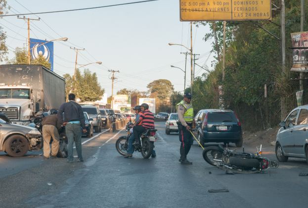 Eine Gruppe von Menschen steht um ein verunglücktes Motorrad auf dem Seitenstreifen, im Hintergrund sind mehrere Fahrzeuge, darunter ein Lkw, sowie Bäume, Pfosten, Laternen, Schilder und der Himmel zu sehen.