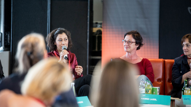 Eine Gruppe von Frauen sitzt vorne, wobei eine links eine rote Kleidung trägt und ein Mikrofon hält, und ein rollbares Banner und eine schwarze Folie im Hintergrund.