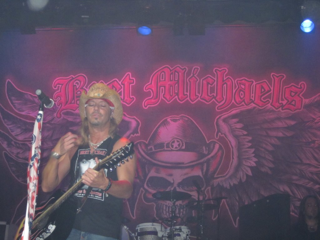 Eine Musik-Konzert-Szene mit Scheinwerfern oben, einem Banner im Hintergrund, Drogen in der Mitte unten, einer Person in der rechten unteren Ecke und einem Gitarristen mit Mikrofon links.