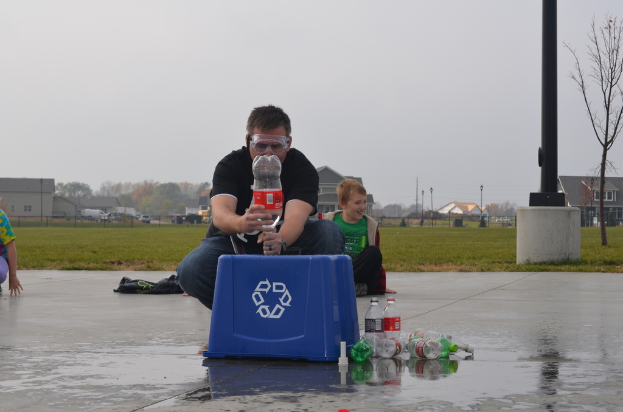 Ein Mann mit Brille hält eine Flasche und steht zwischen mehreren Flaschen und einer Box, umgeben von Pfählen, Bäumen, Gebäuden, Fahrzeugen und Gras.