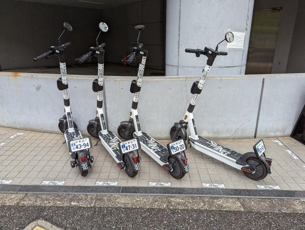 Drei schwarze und weiße E-Scooter mit Nummern und Text nebeneinander auf einem Gehweg vor einem Gebäude mit einer Wand und einer Glastür im Hintergrund geparkt.