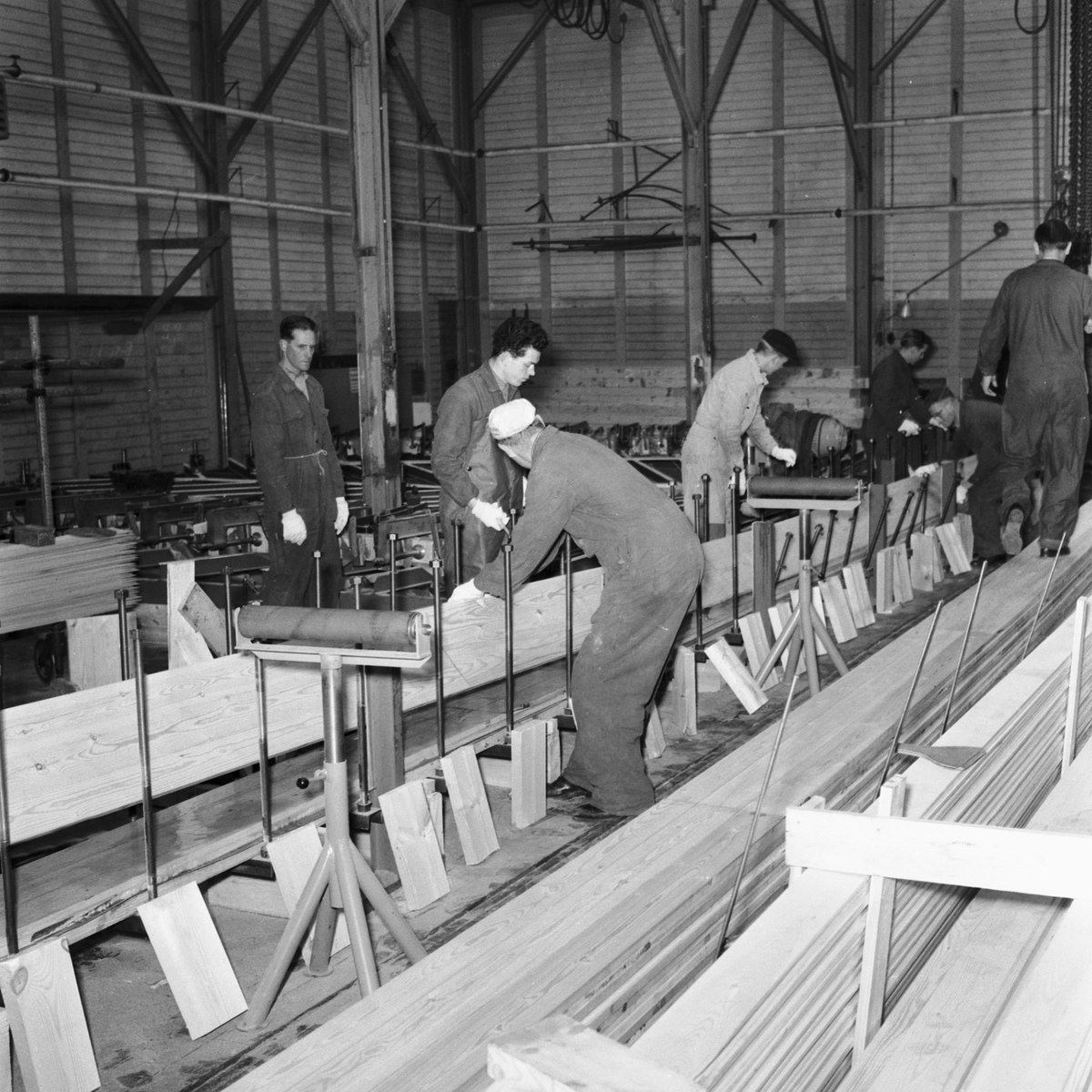 Schwarzes und weißes Bild von Männern, die in einer Fabrik mit Holzplanken auf dem Boden und Maschinen im Hintergrund arbeiten.
