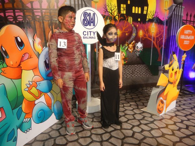 Zwei Kinder in Halloween-Kostümen stehen nebeneinander vor einer Wand mit einer Painting eines Gebäudes, Bäumen, Laternenmasten und einem Zaun, mit Schildern mit Text und Tierbildern.