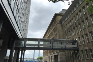 Ein Auto fährt eine Straße mit hohen Gebäuden entlang, mit einer Fußgängerbrücke in der Mitte, Menschen auf dem Gehweg und Bäumen, Pfählen und einem bewölkten Himmel im Hintergrund.