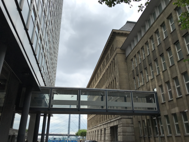 Ein Auto fährt eine Straße mit hohen Gebäuden entlang, mit einer Fußgängerbrücke in der Mitte, Menschen auf dem Gehweg und Bäumen, Pfählen und einem bewölkten Himmel im Hintergrund.
